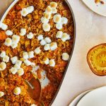 Mashed sweet potatoes with marshmallows on top and a golden spoon in a white casserole dish.