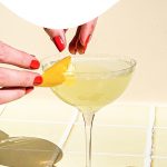 two hands placing lemon peel on lemon martini in coupe glass