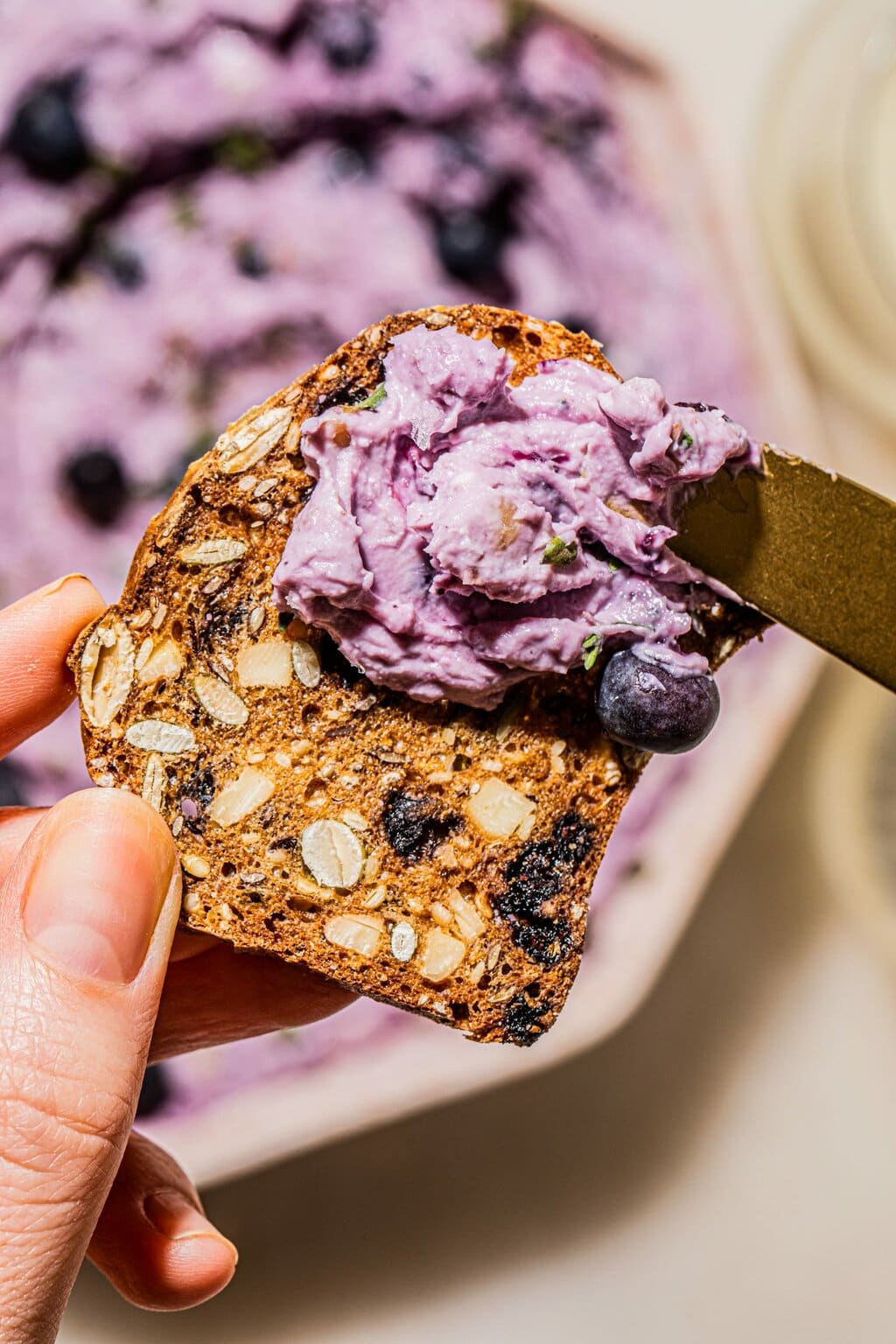 two hands spreading goat cheese with blueberries on cracker with gold knife