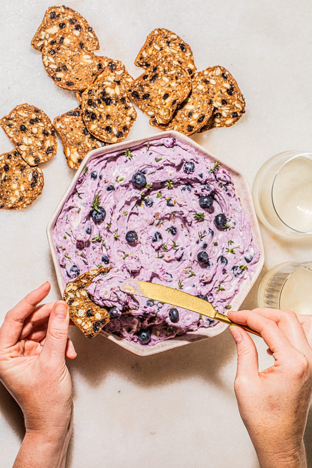 two hands spreading blueberry goat cheese on cracker with gold knife