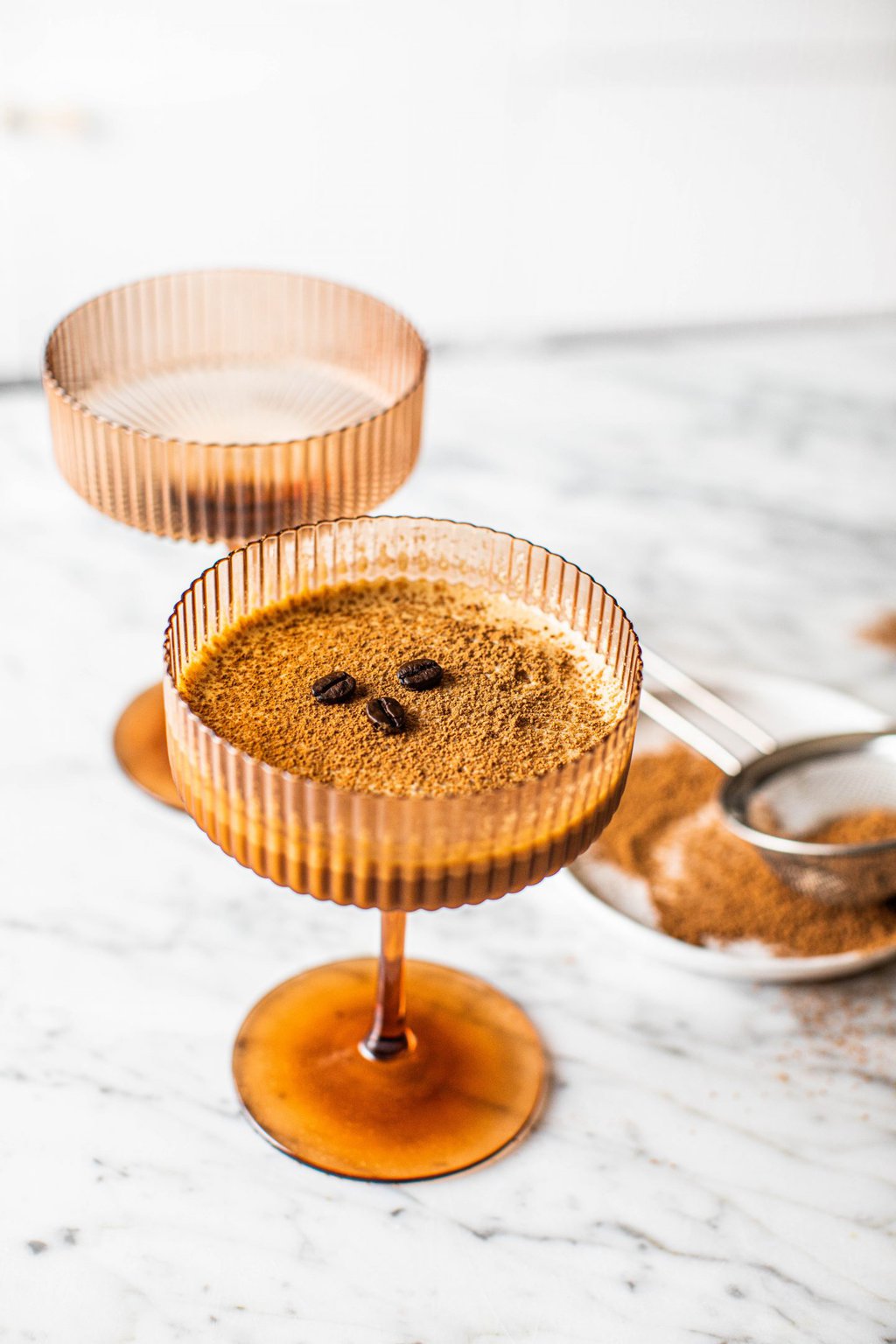 a brown coupe glass filled with a non alcoholic espresso martini topped with espresso beans and sifted with cocoa powder 