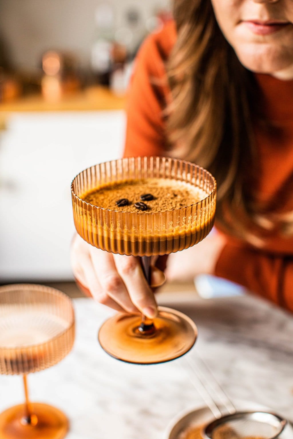  a girl in an orange shirt taking a sip of a non alcoholic espresso martini in a brown coupe glass 