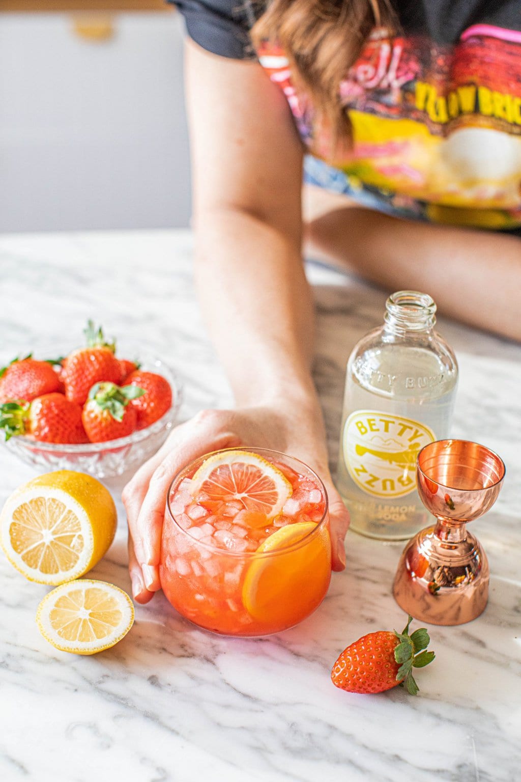 a hand grabbing a strawberry drink glass with lemon slice, strawberries, lemon soda and copper jigger 