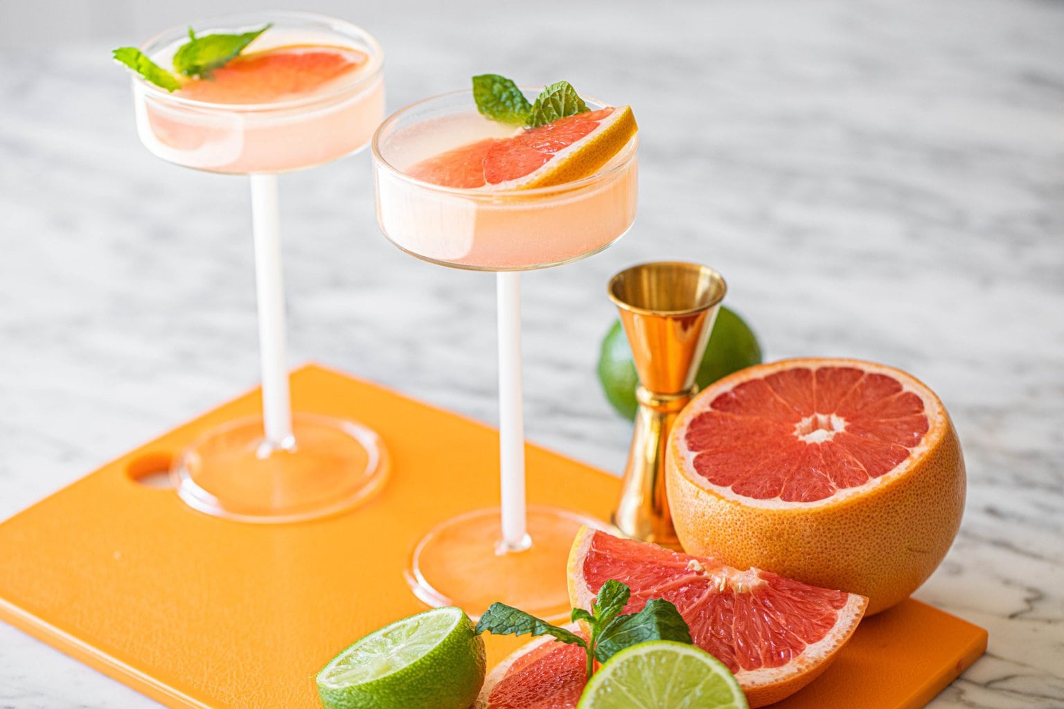 two ruby red grapefruit martinis in coupe glasses on orange board surrounded by slices of grapefruits and limes