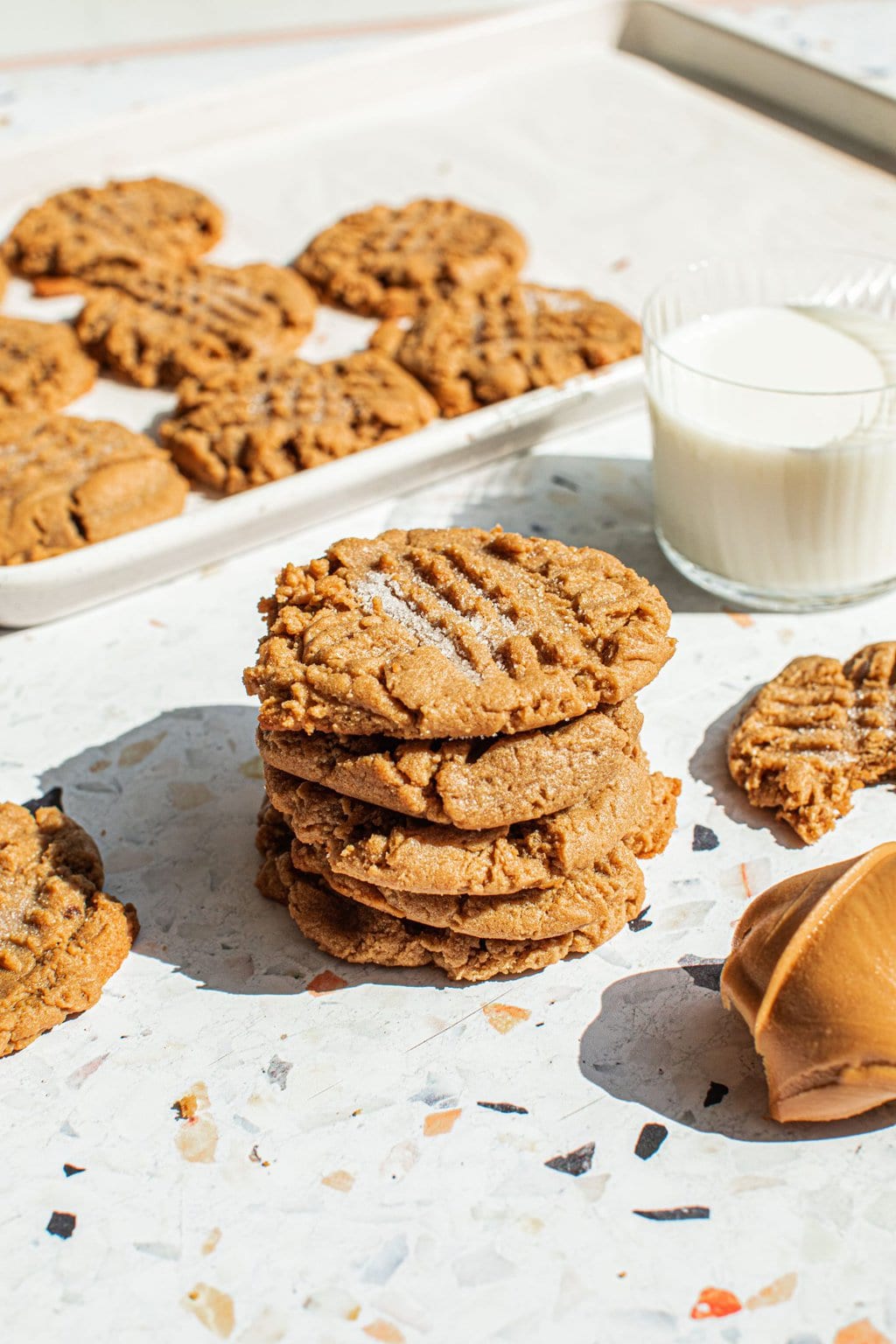 a stack of peanut butter cookies with a tray of more cookies behind them along with a glass of milk