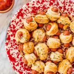 a red and white plate full of air fried pigs in a blanket with two small bowls of ketchup and honey mustard in the upper left corner