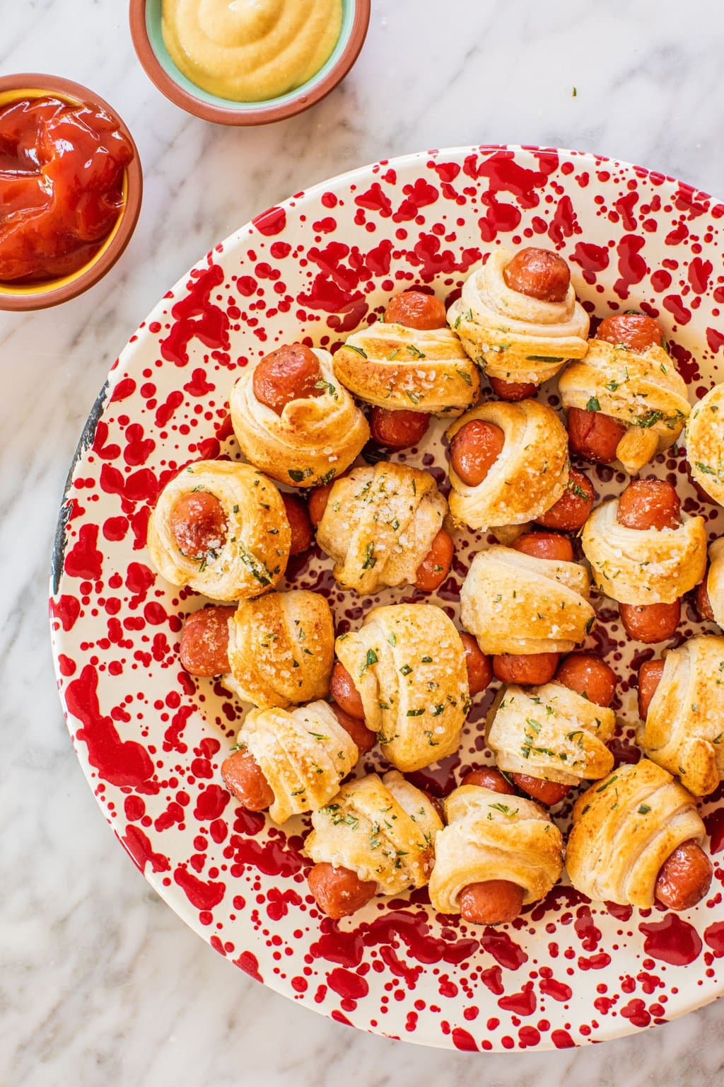 a red and white plate full of air fried pigs in a blanket with two small bowls of ketchup and honey mustard in the upper left corner