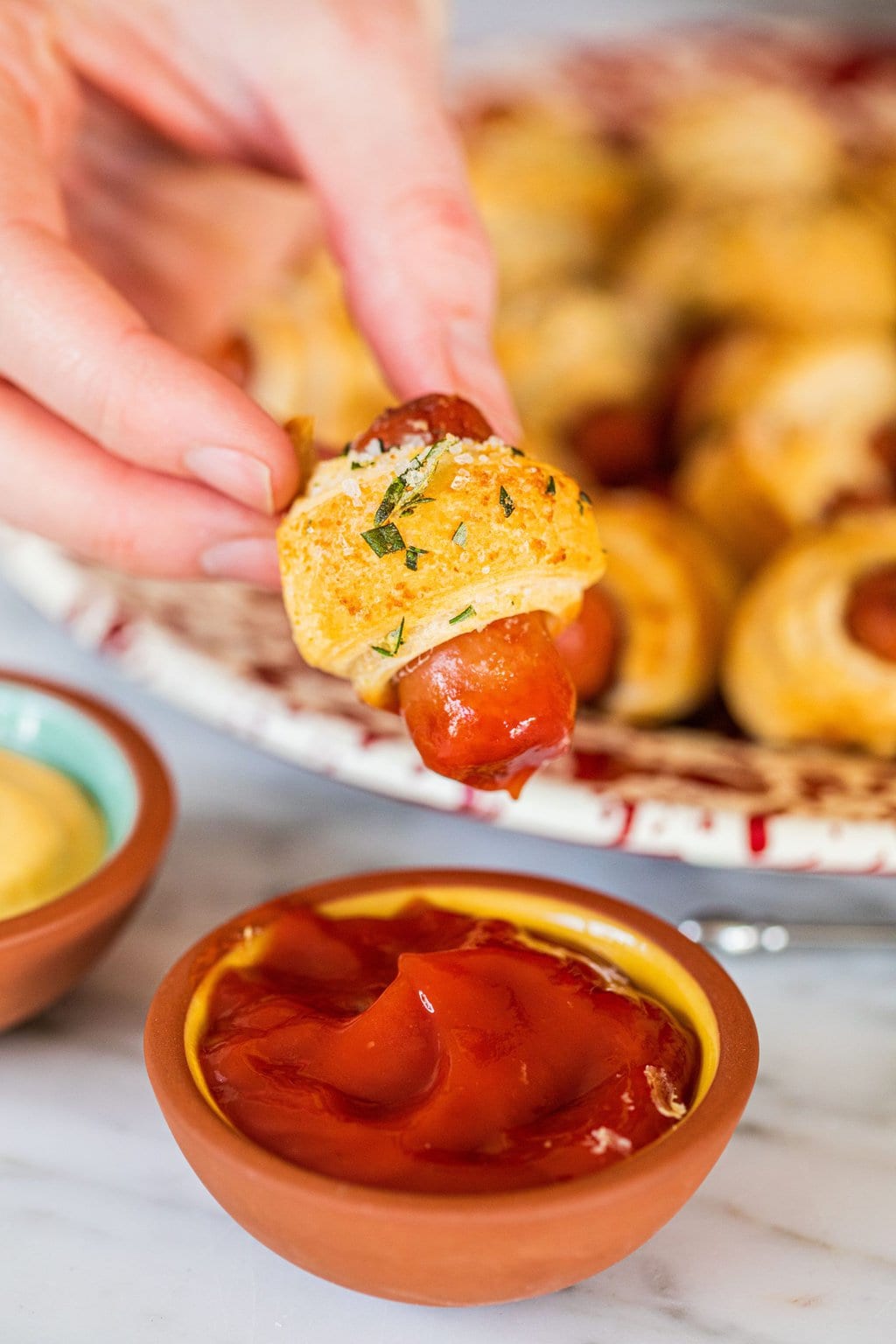 a single dough wrapped hot dog being dipped in a small bowl of ketchup with other hot dogs out of focus on a plate in the background