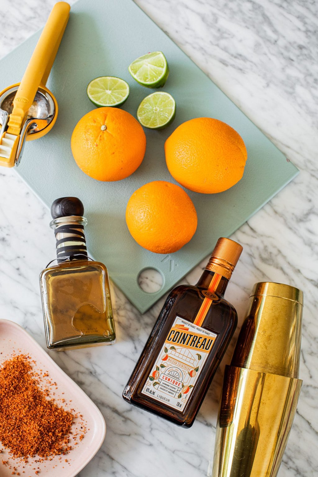 oranges, tequila, cointreau, limes, cara cara oranges, tajin, and a cocktail shaker on a white marble countertop and a light blue cutting board