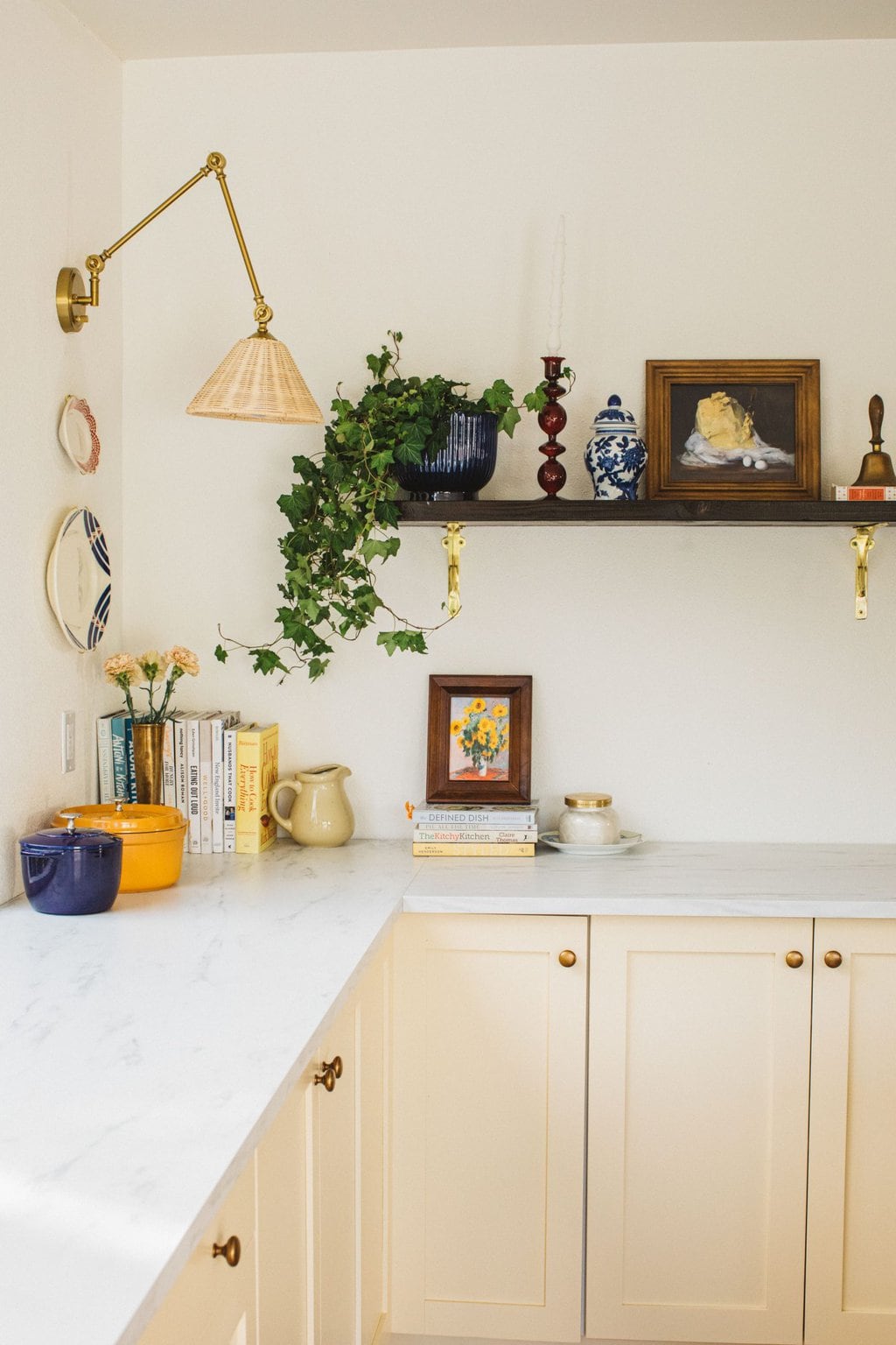 a pool house kitchen with a wicker brass sconce, open shelving, butter print, large ivy plant and cream cabinets 