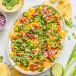 a bowl of five layer bean dip with chips, limes, green onion, jalepenos, etc scattered around it