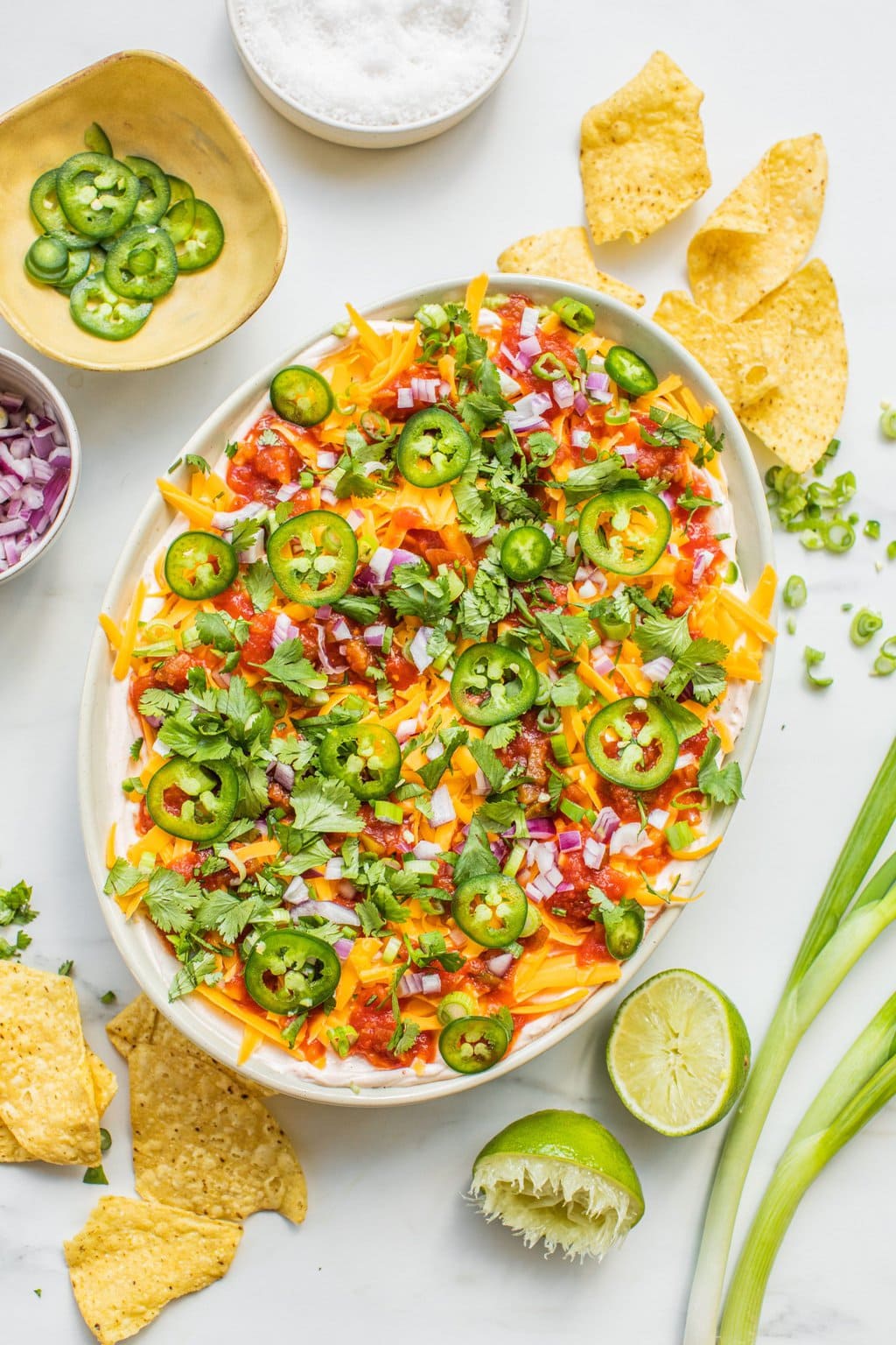 a bowl of five layer bean dip with chips, limes, green onion, jalepenos, etc scattered around it