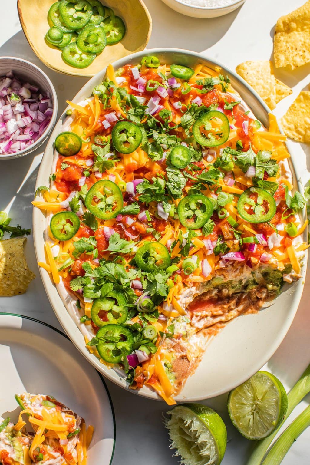 a large white bowl full of bean dip with a serving missing in the bottom right corner, and various ingredients and chips scattered around