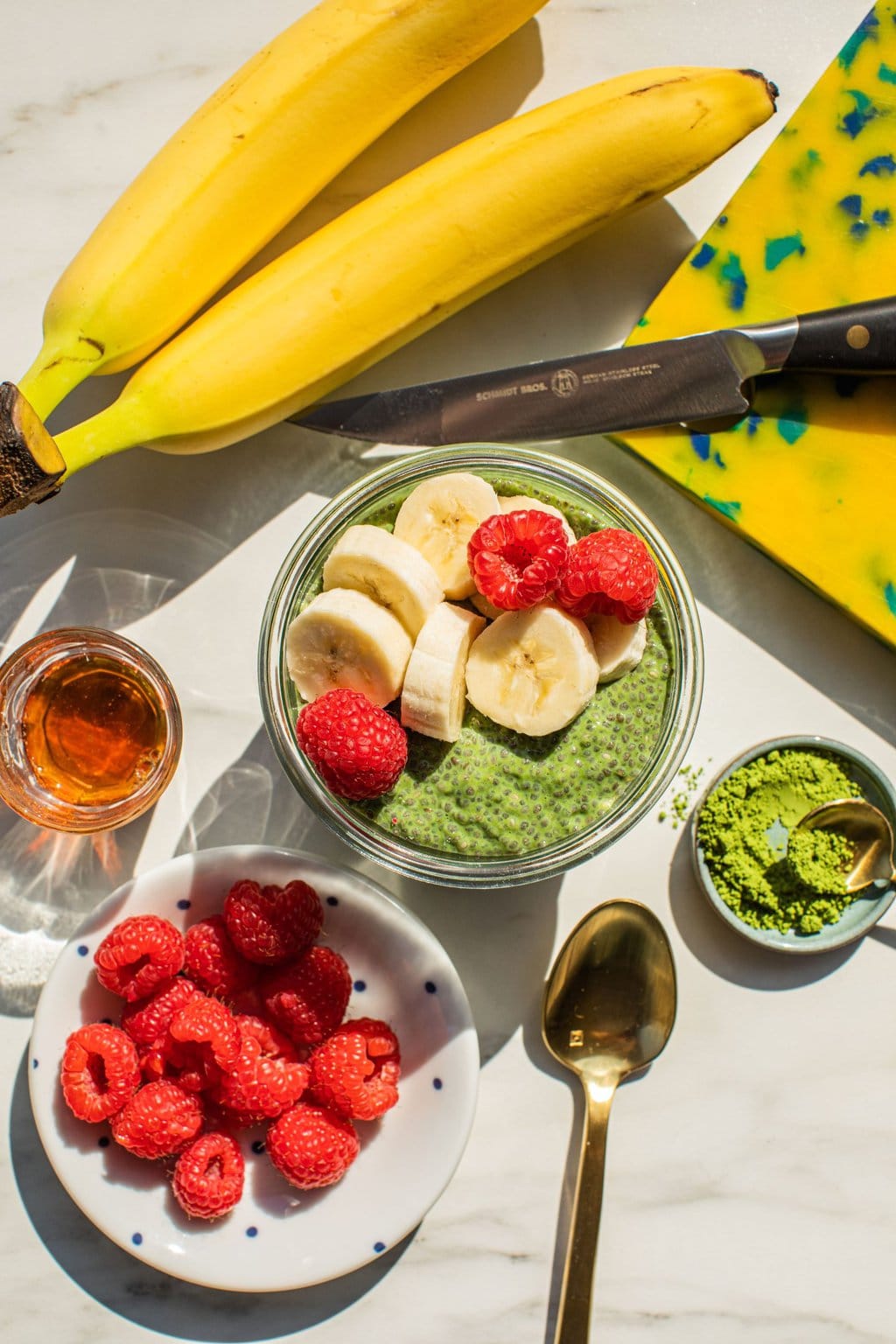 a bowl of matcha chia seed pudding with bananas and raspberries on top with the ingredients surrounding the bowl