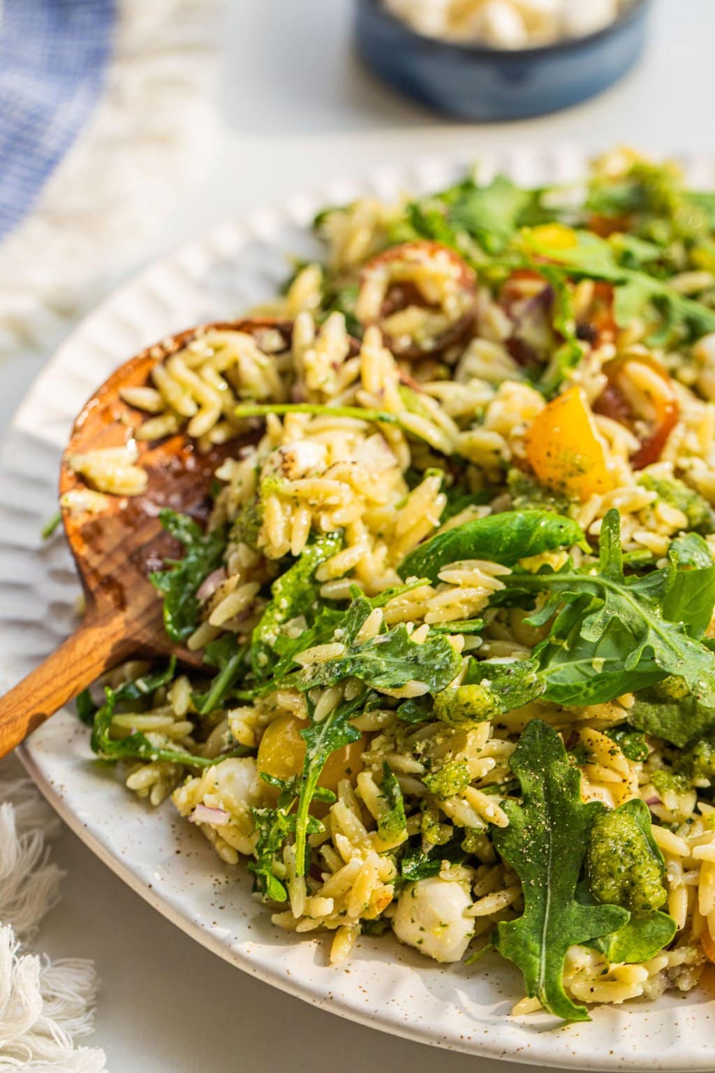 a wooden spoon sticking out of a serving of pesto orzo salad with arugula, cherry tomatoes, and mozzarella