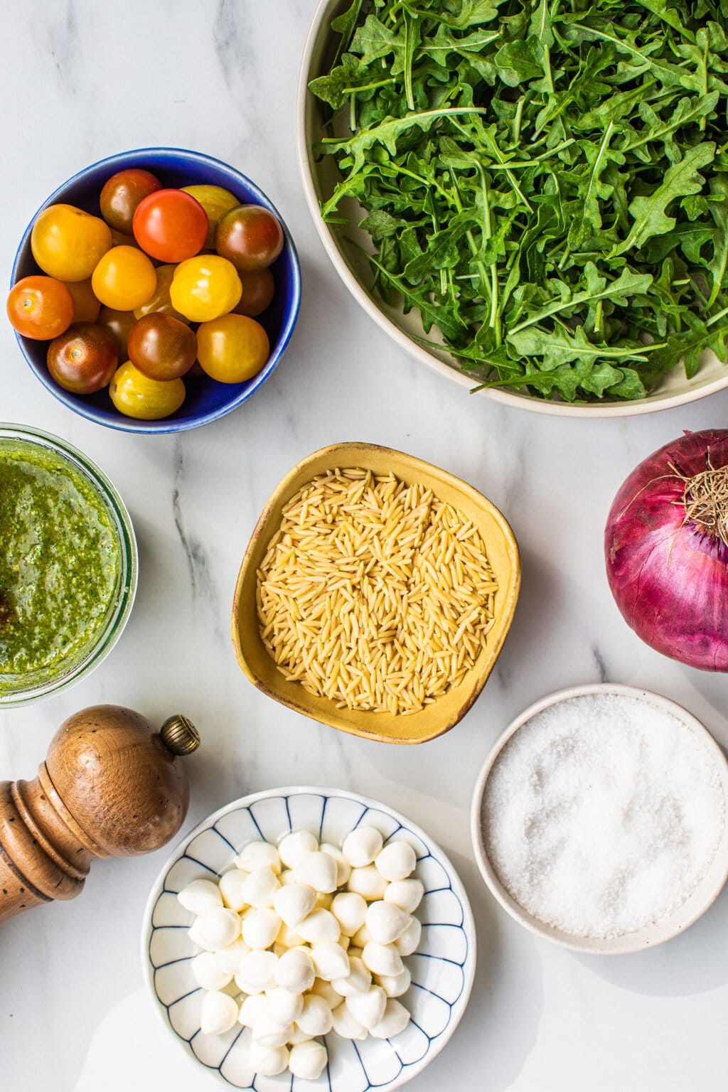 bowls of orzo, cherry tomatoes, mozarella pearls, pesto, salt, arugula, and a whole red onion