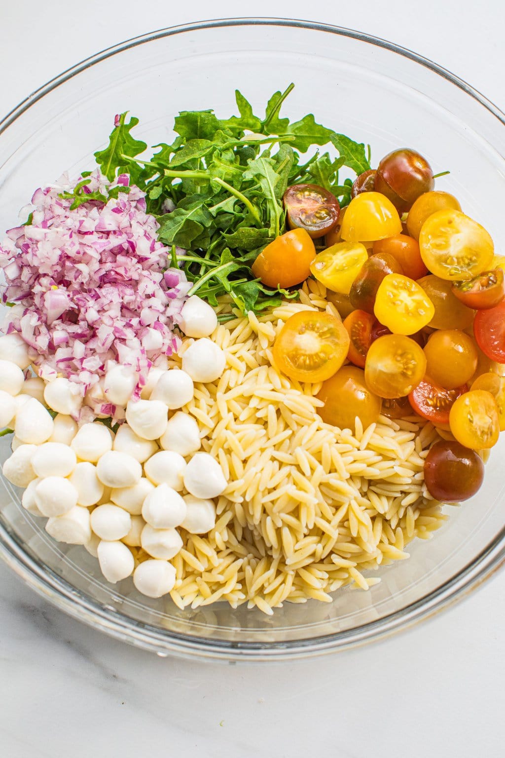 a white bowl full of sections of chopped red onion, mozzarella pearls, cooked orzo, sliced cherry tomatoes, and chopped arugula