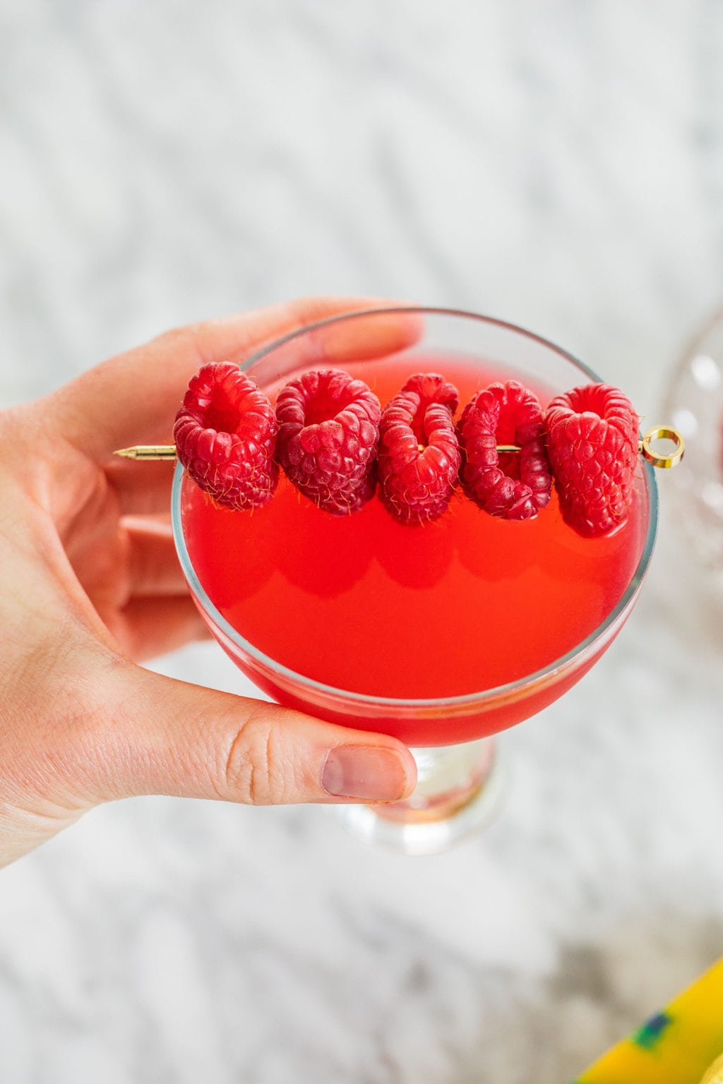 a red martini held by elizabeth with five raspberries garnishing skewered by a decorative toothpick as garnish