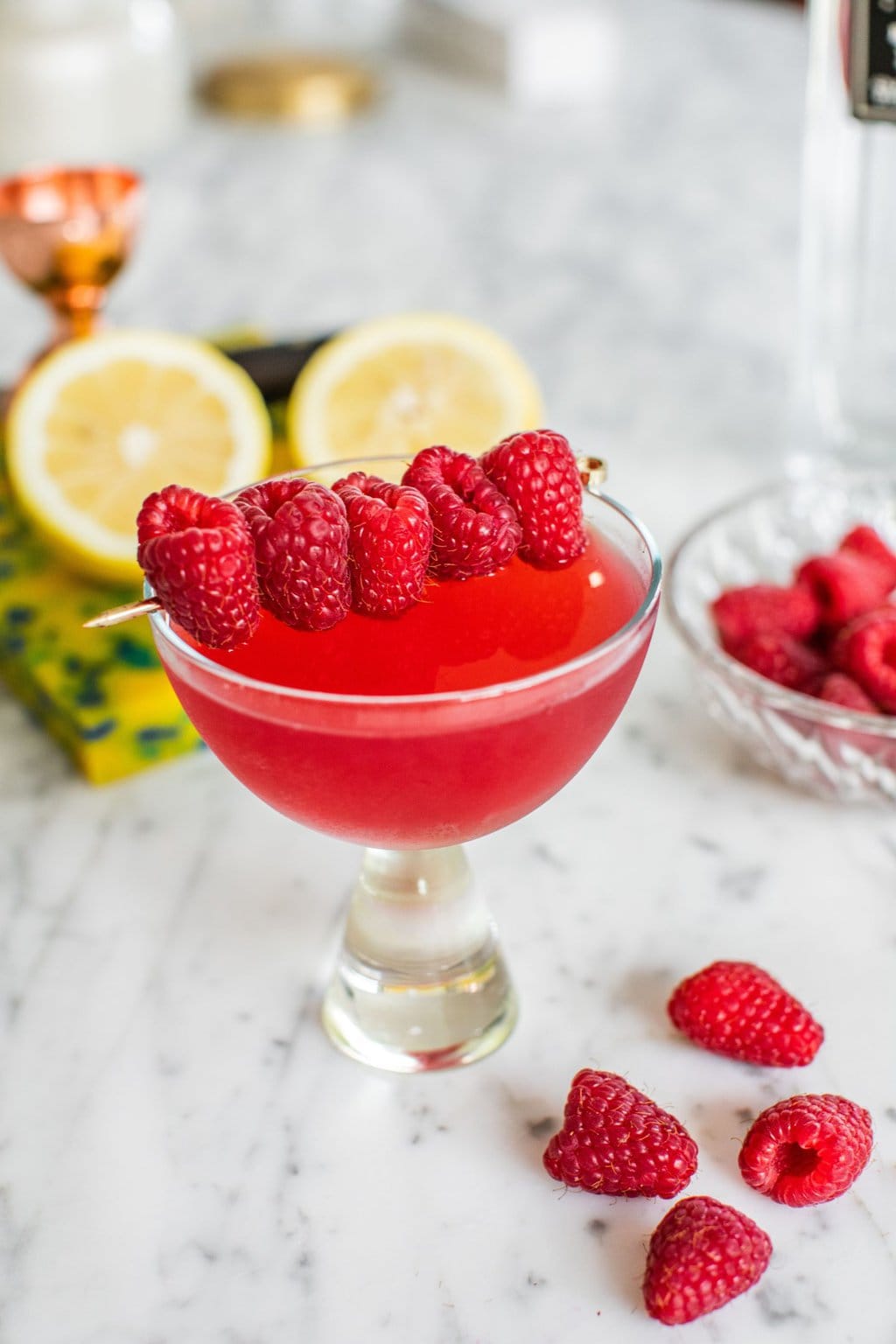 a raspberry martini on a white marble counter top garnished with a toothpick stacked with raspberries