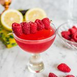 a raspberry martini on a white marble counter top garnished with a toothpick stacked with raspberries