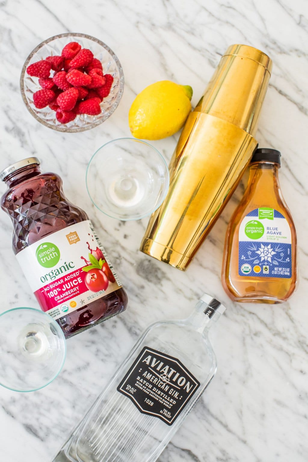 alcohol, agave, cocktail shaker, cranberry juice, lemon, and raspberries on a marble counter top