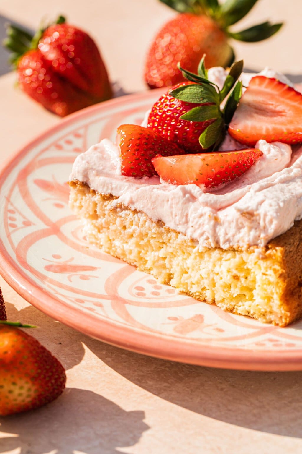 a slice of strawberry tres leches cake on a decorative pink plate with strawberries on top and scattered around the cake