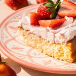 a slice of strawberry tres leches cake on a decorative pink plate with strawberries on top and scattered around the cake