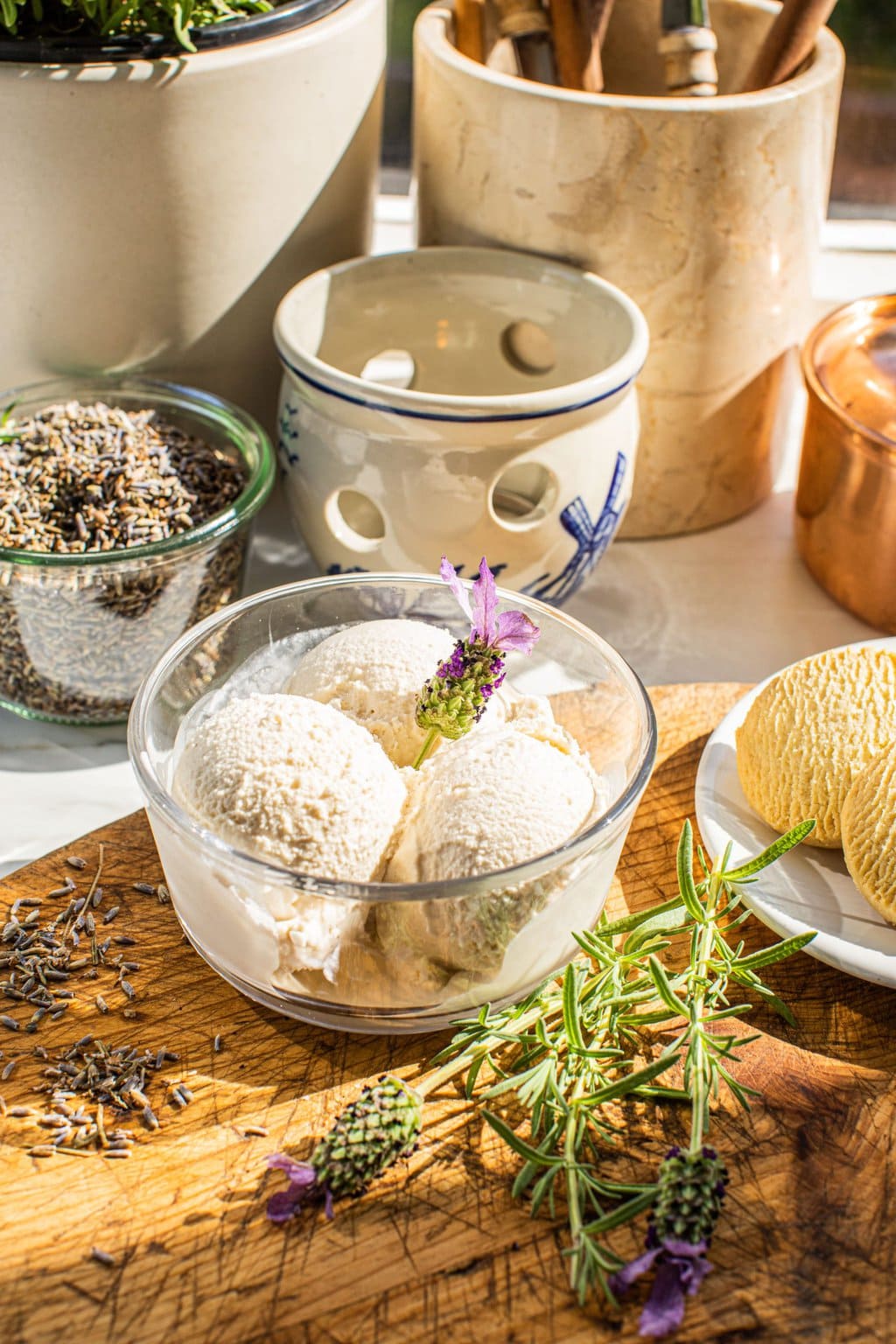 a glass of three scoops of lavender ice cream sitting on a brown chopping board that has lavender scattered around and cooking utensils in the background