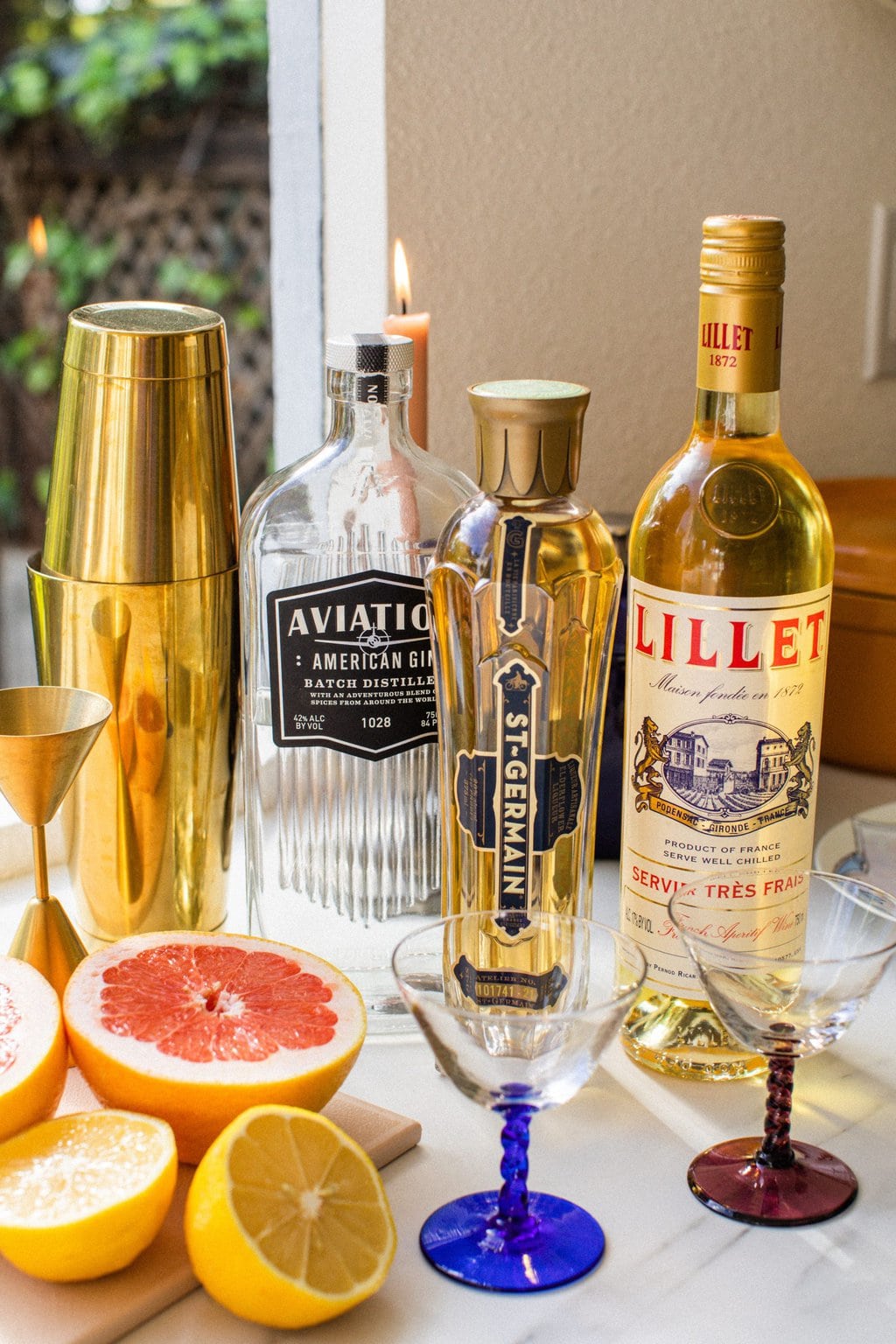 a gold cocktail shaker, a bottle of aviation gin, a bottle of St. Germain elderflower liqueur, lillet, and slices of grapefruit and lemon beside cocktail glasses