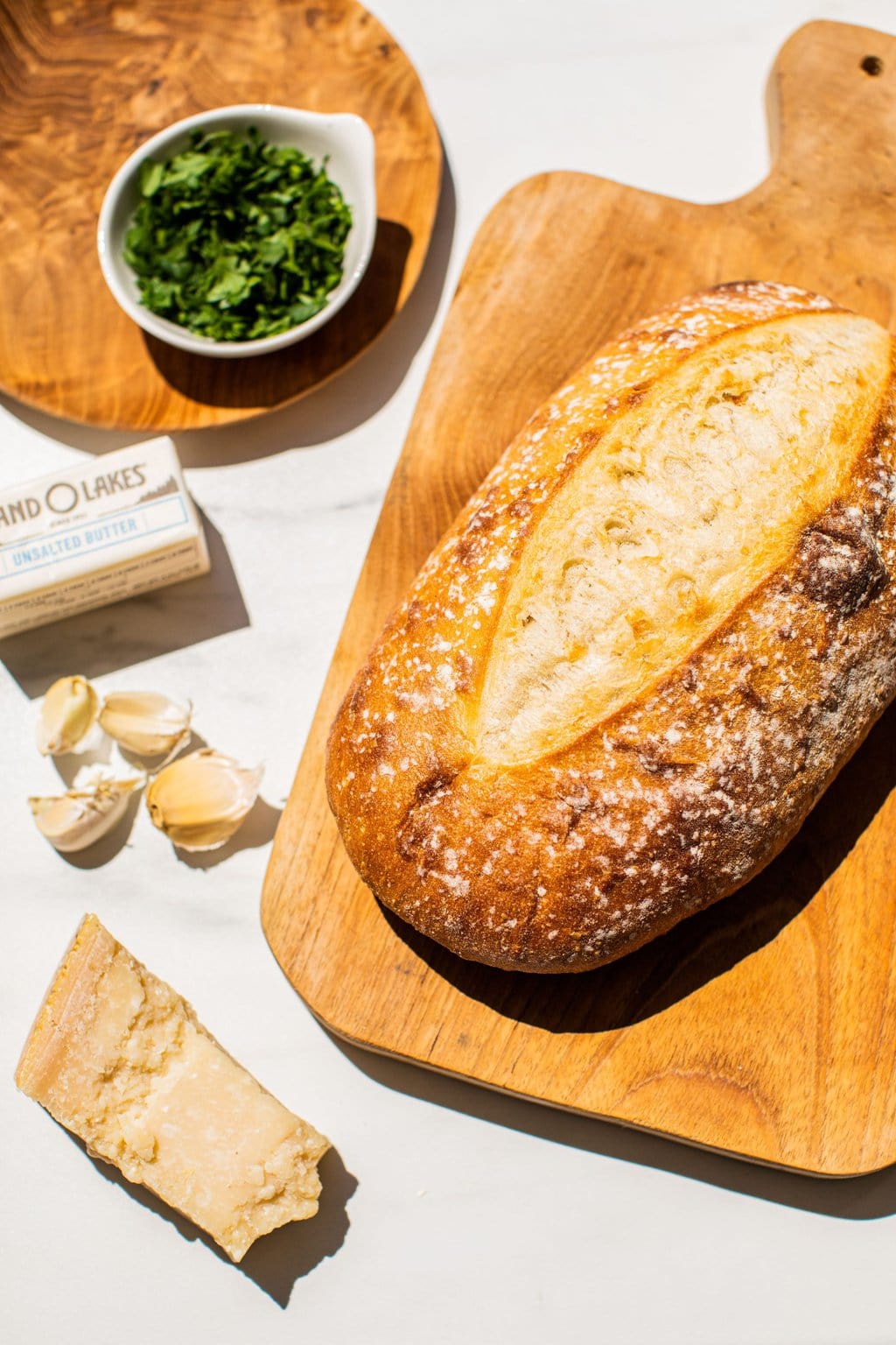 a loaf of french bread on a wooden board with parsley, butter, garlic, and parmesan to the left