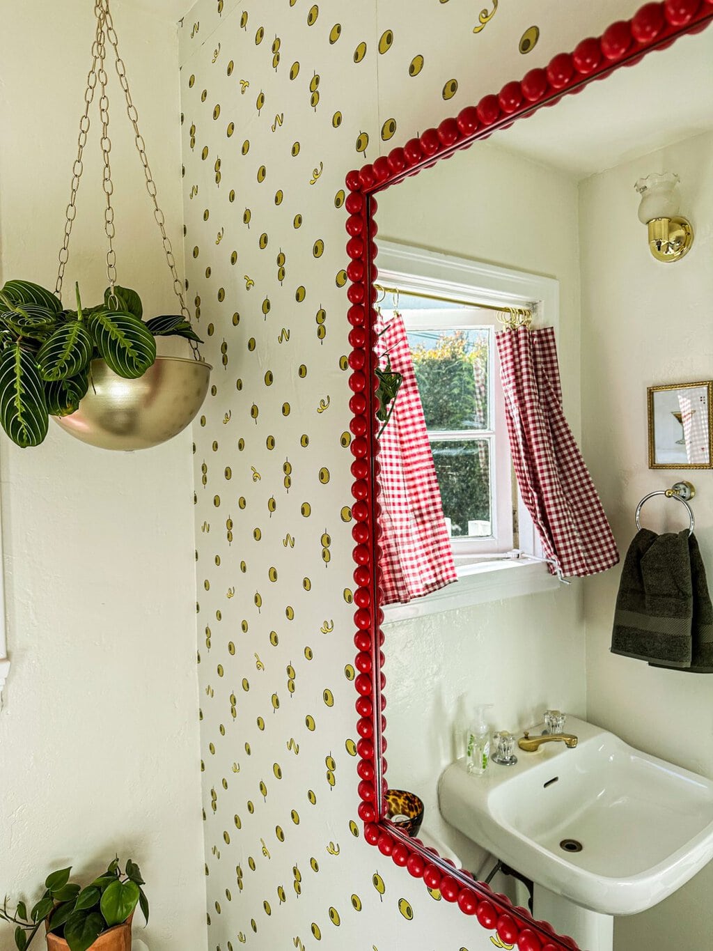 a red bobbin mirror on top of olive and lemon wallpaper with a hanging plant in the left corner. In the mirrors reflection is a window with red checkered curtains and a an olive green hand towel 