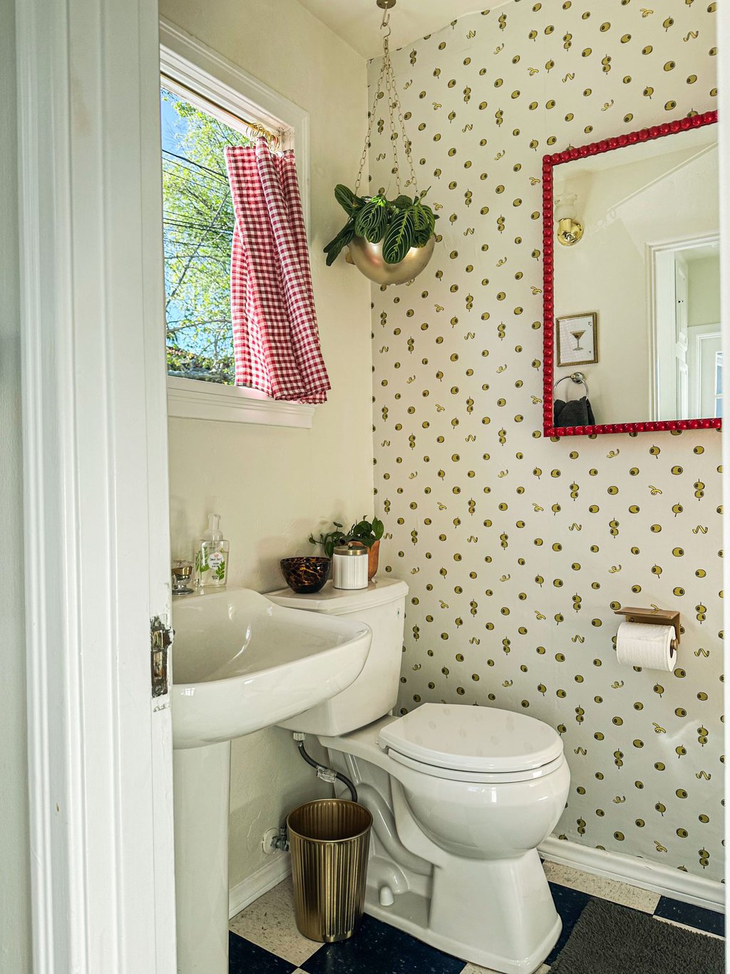 The bathroom with a toilet, pedestal sink, a plant hanging in the corner, a red french bobbin mirror, olive wallpaper, and red checkered drapes over the window