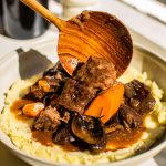 a bowl of beef bourguignon served over mashed potatoes with a wood spoon adding sauce to the dish