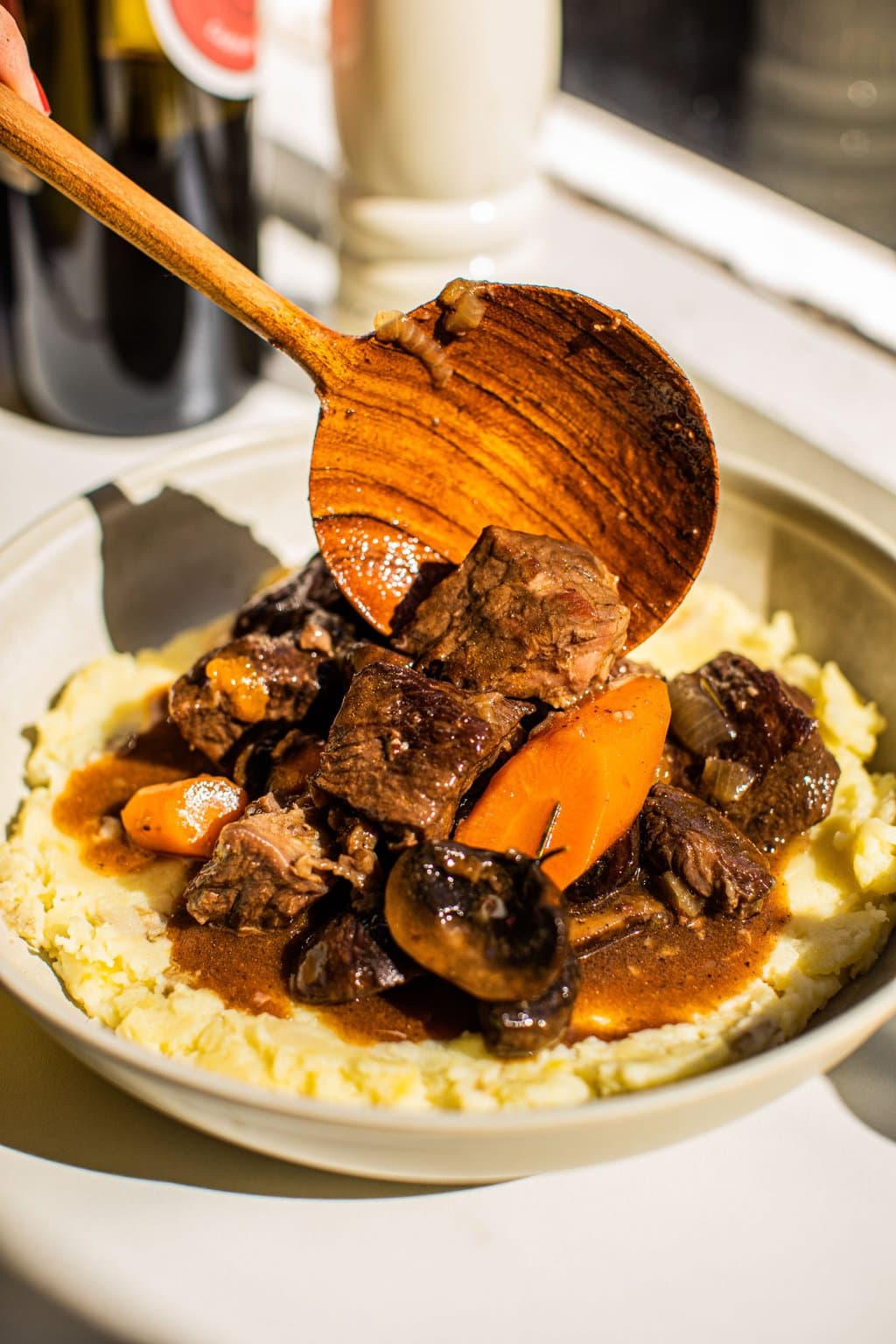 a bowl of beef bourguignon served over mashed potatoes with a wood spoon adding sauce to the dish