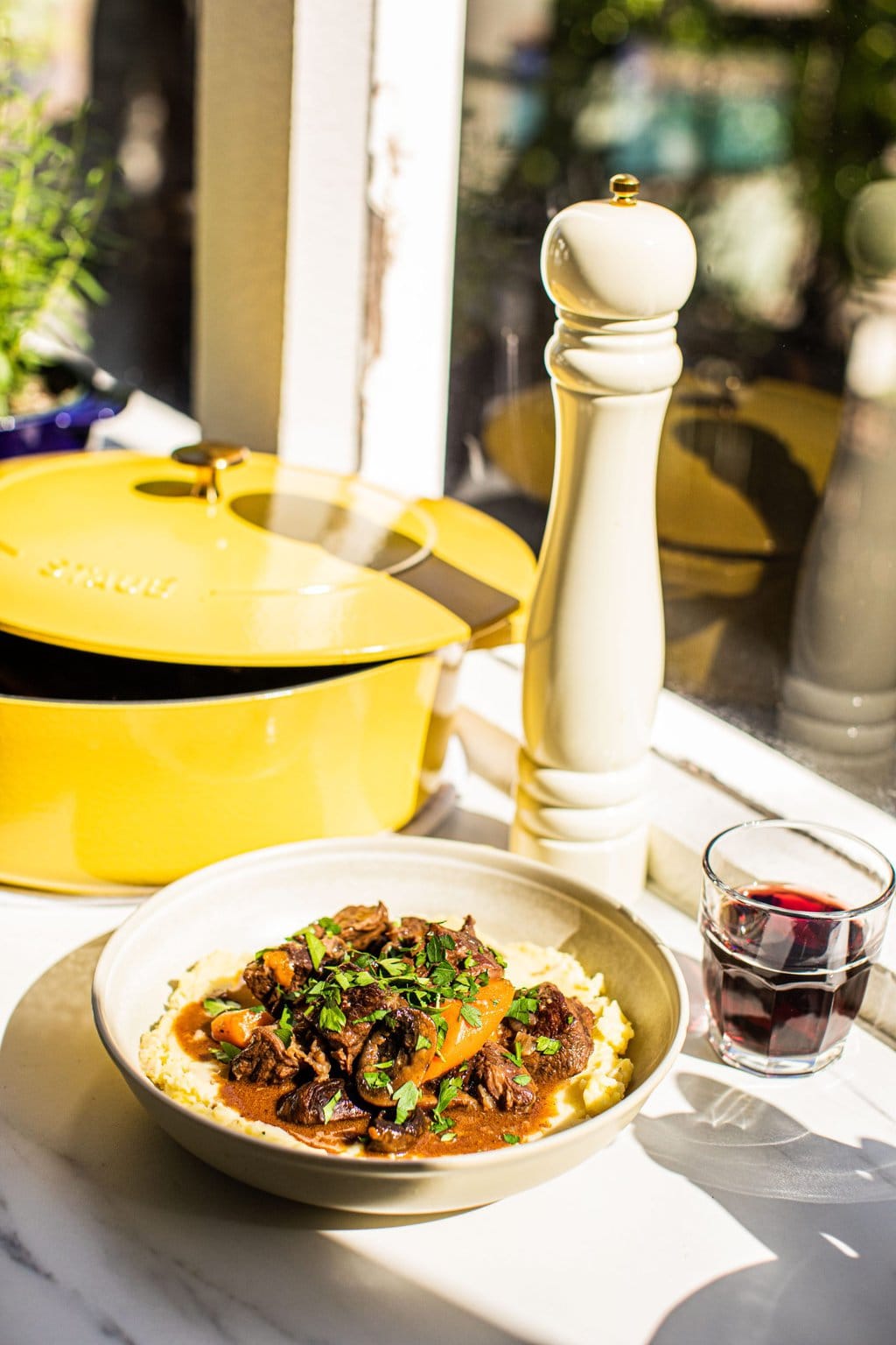 a bowl of red wine braised beef with veggies served over mashed potatoes alongside a glass of wine