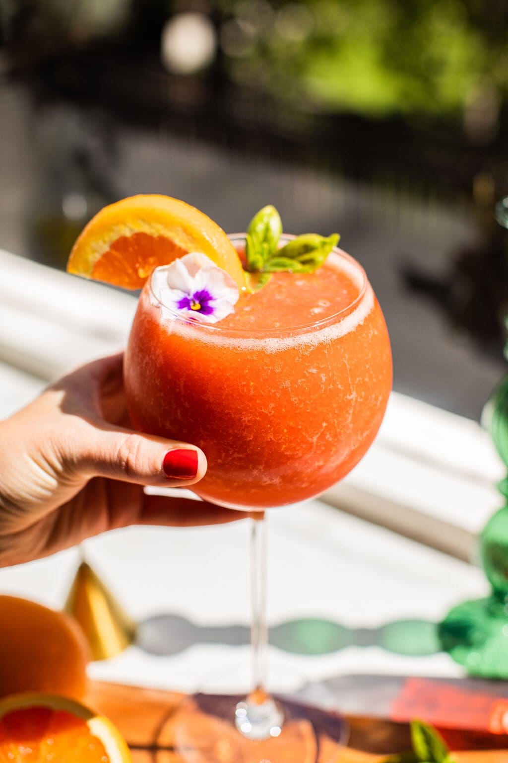 elizabeth holding a glass of frozen strawberry spritz with an orange slice and flower garnish on top