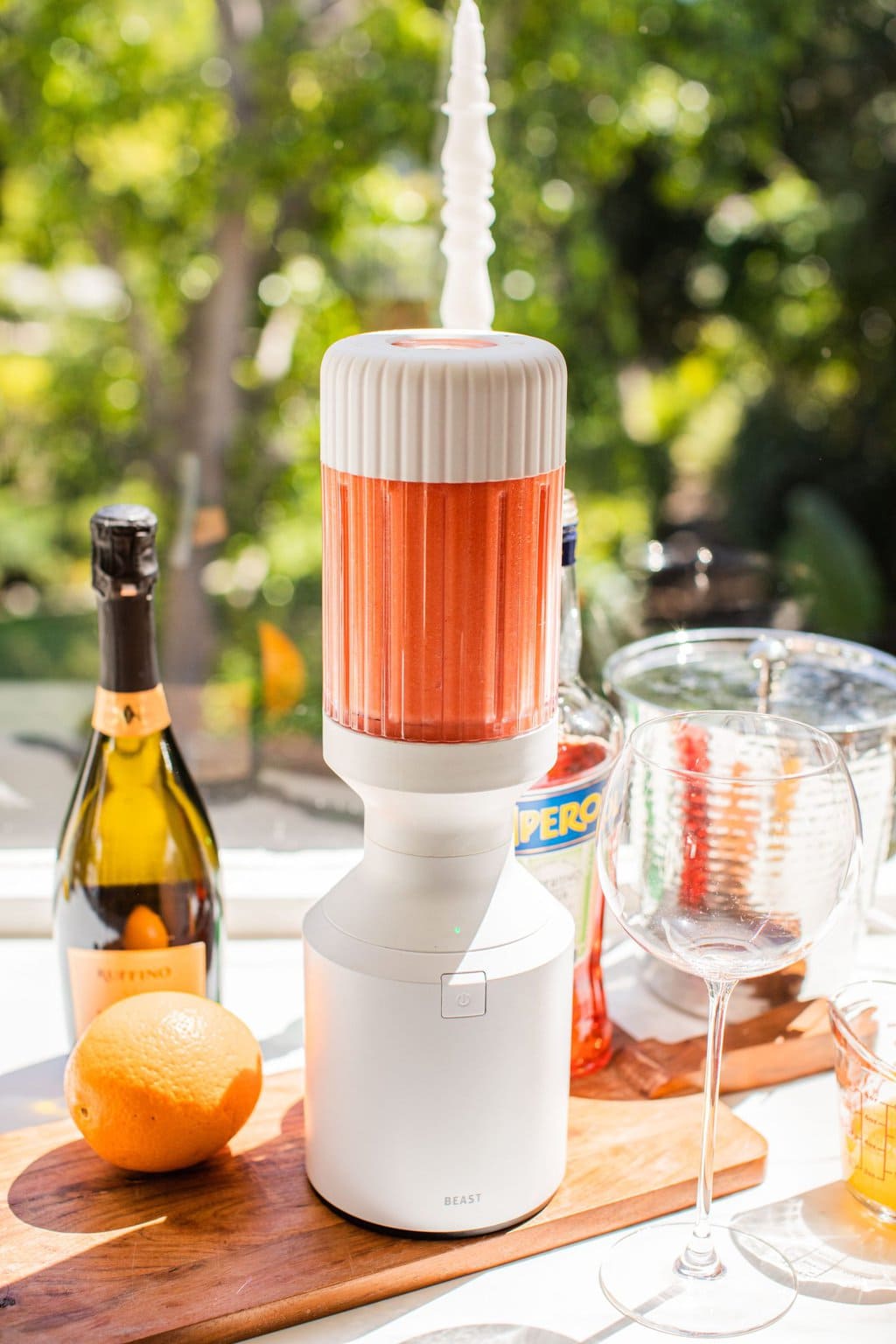 a blender full of frozen spritz beside a glass, an orange, some prosecco, and aperol in the background