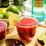 cherry margarita garnished with lime, with a bottle of italicus in the background and a bowl of cherries in the foreground