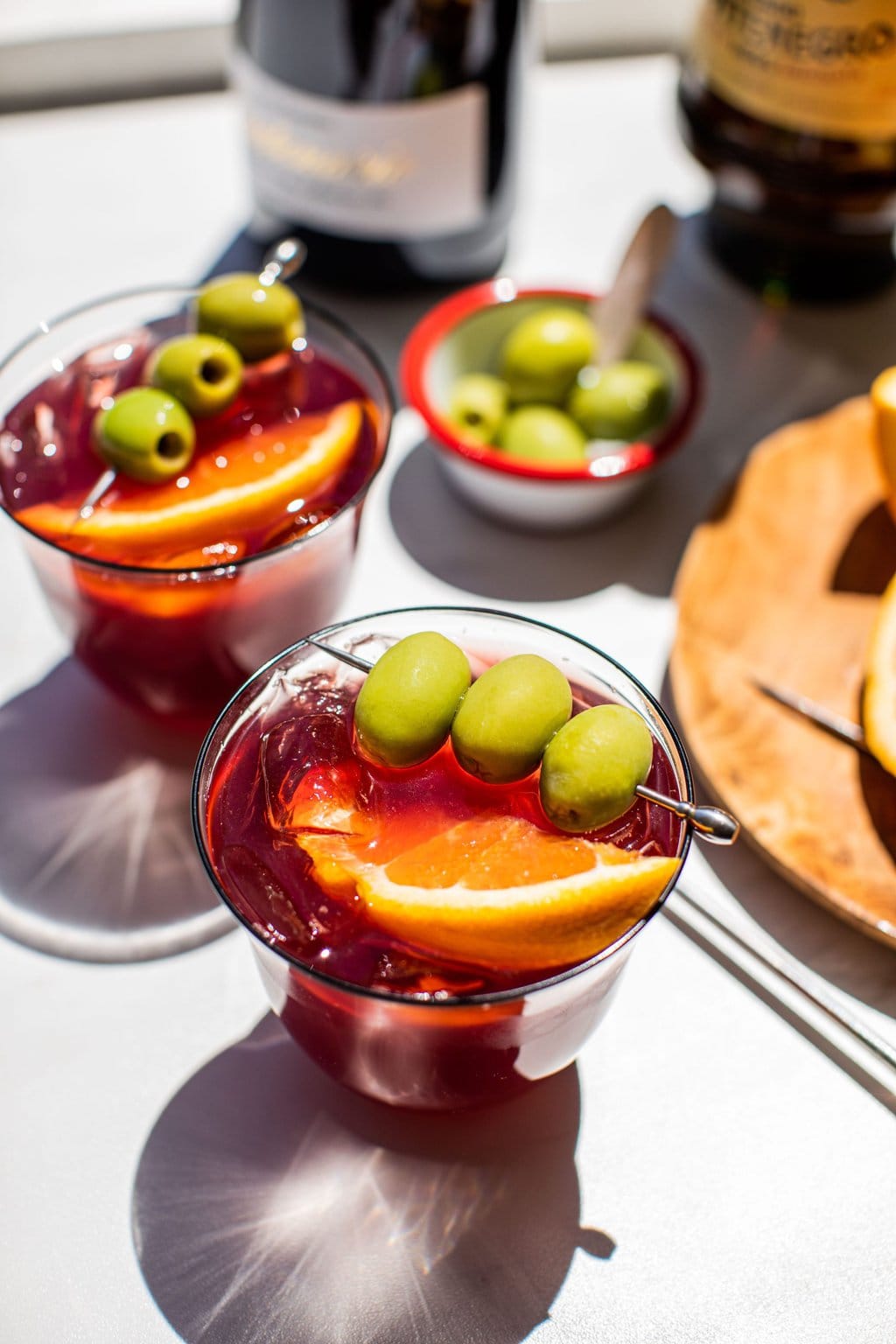 two low ball glasses filled with lambrusco spritz and garnished with a silver cocktail pick with three green olives and a large citrus wedge. Small bowl of green olives in the back. 