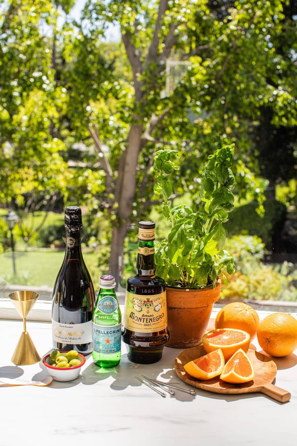 a gold jigger, small white bowl with green olives, a bottle of lambrusco sparkling red wine, a small green bottle of pellegrino, a large bottle of Amaro, a basil plant in a terra cotta pot, a few citrus wedges on a small wooden cutting board 