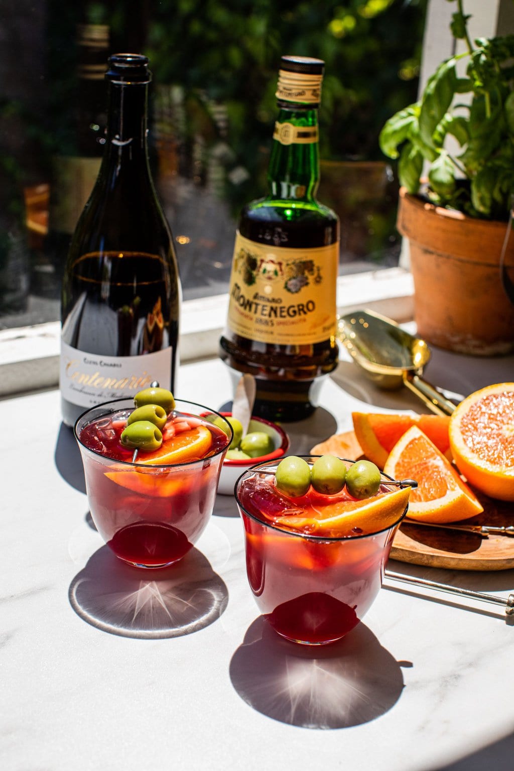 two lambrusco spritz glasses filled with silver cocktail picks with three green olives and a large citrus wedge. in the back is a bottle of lambrusco and a bottle of amaro. Citrus wedges sitting on top of a small wooden cutting board. 