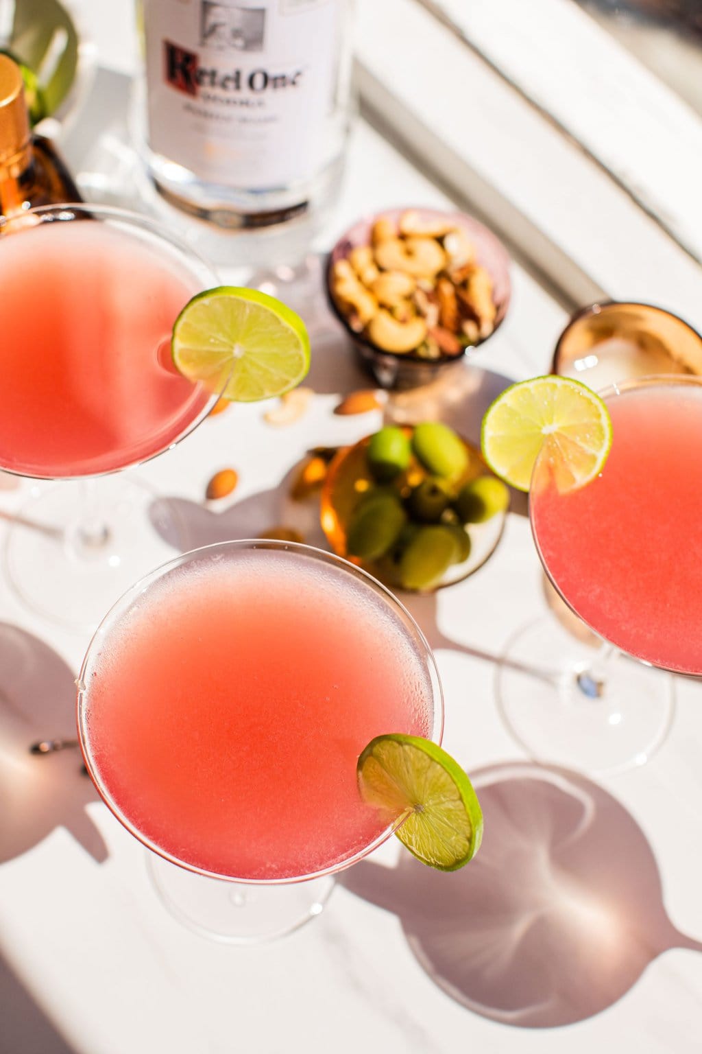 three cosmopolitans garnished with lime photographed from above with nuts, olives, and ketel one pictured in the background