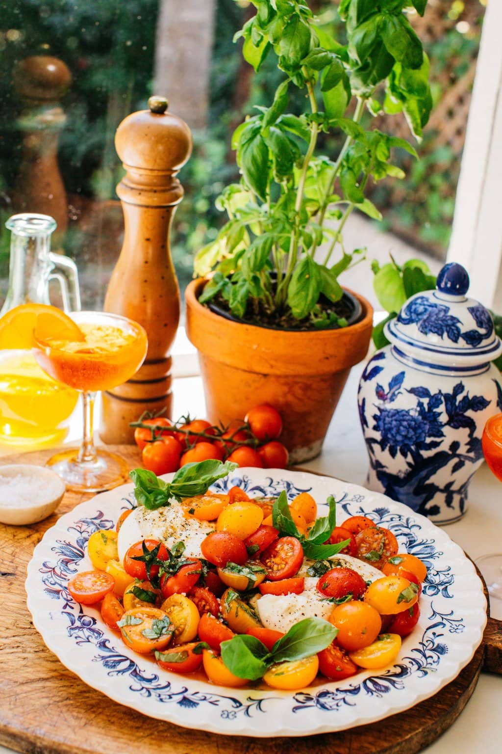 a bowl of caprese salad with a drink, basil plant, pepper, and some other ingredients in the background