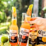 a hand holding a bottle of corona rimmed with tajin and garnished with a lime wedge beside another beer cocktail