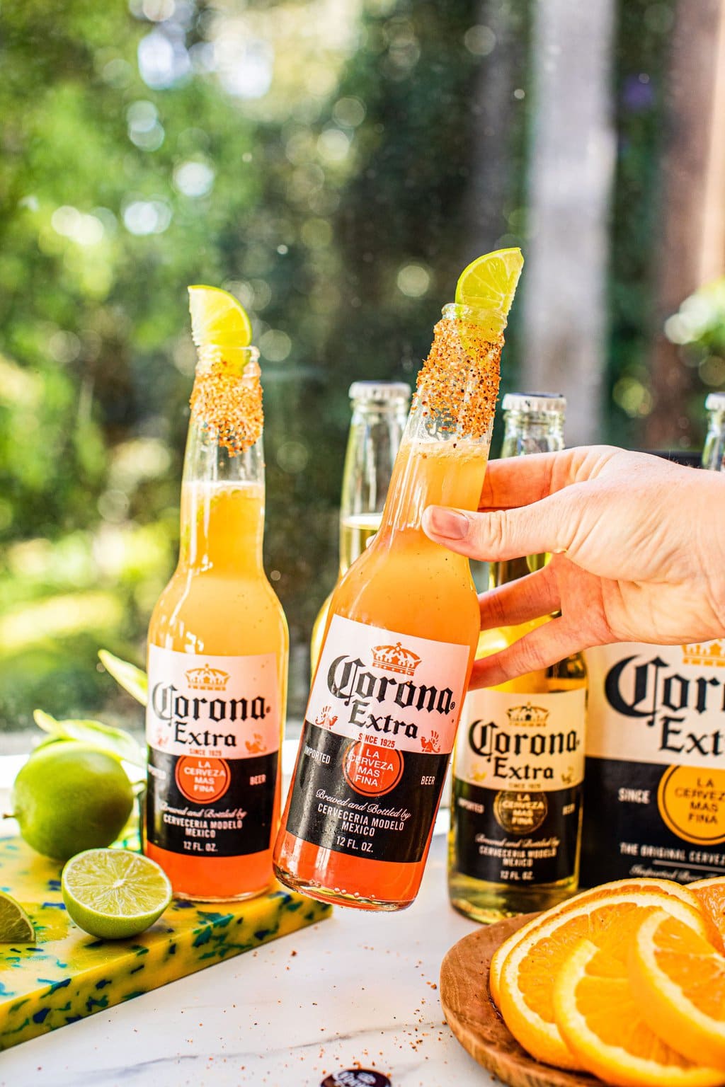 a hand holding a bottle of corona rimmed with tajin and garnished with a lime wedge beside another beer cocktail