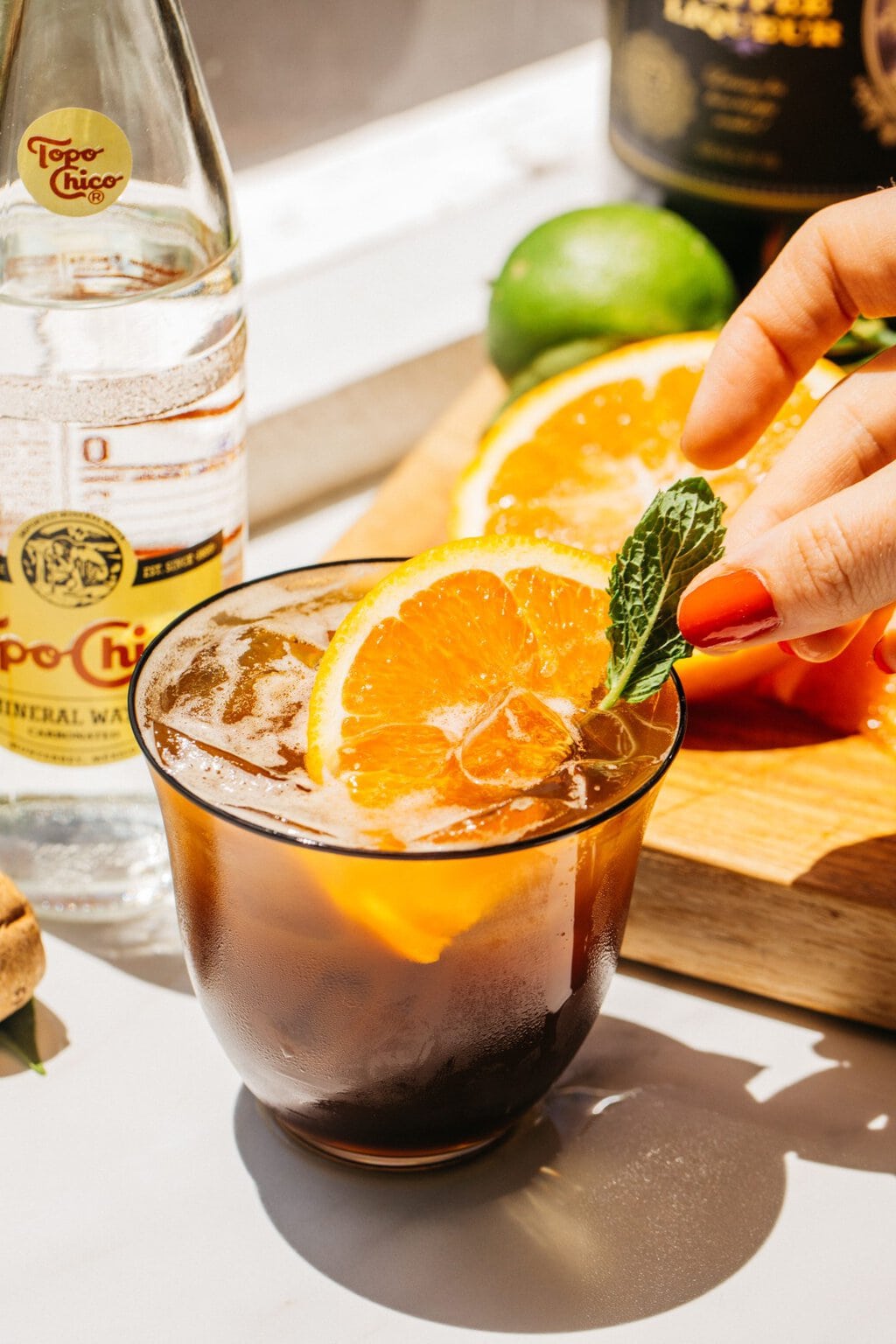 a glass of an espresso spritz with an orange slice for garnish and elizabeth's hand adding a fresh mint leaf