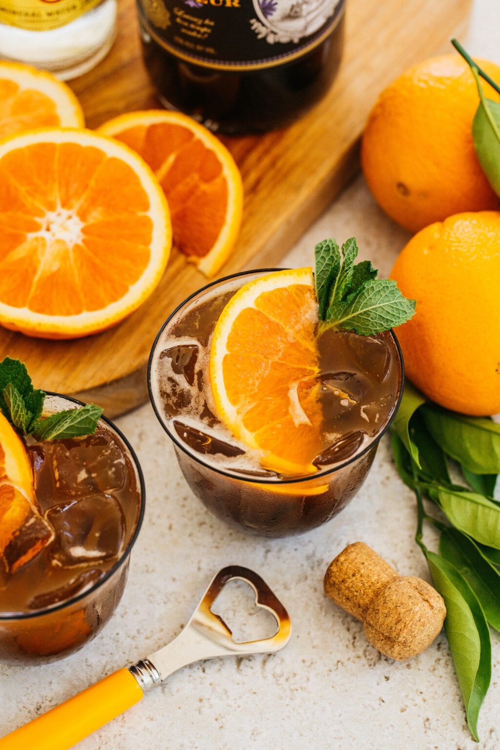 a spritz made with espresso, prosecco, and garnished with a wedge of orange. the drink is surrounded with oranges, another drink, a cork, and a bottle opener