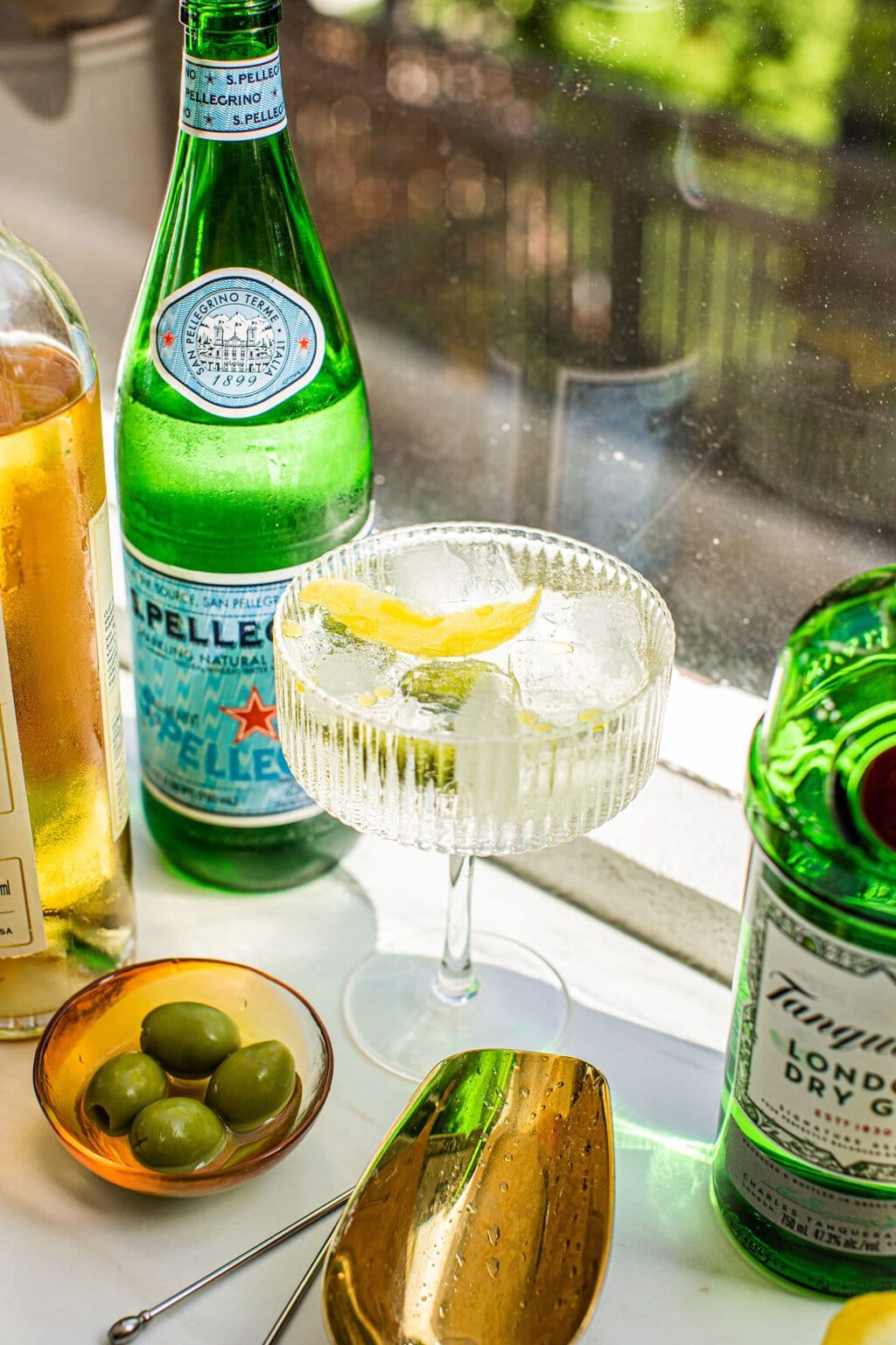 a glass of a spritz with lemon rind and olives in it, surrounded by a bowl of lives, pellegrino, gin, and prosecco