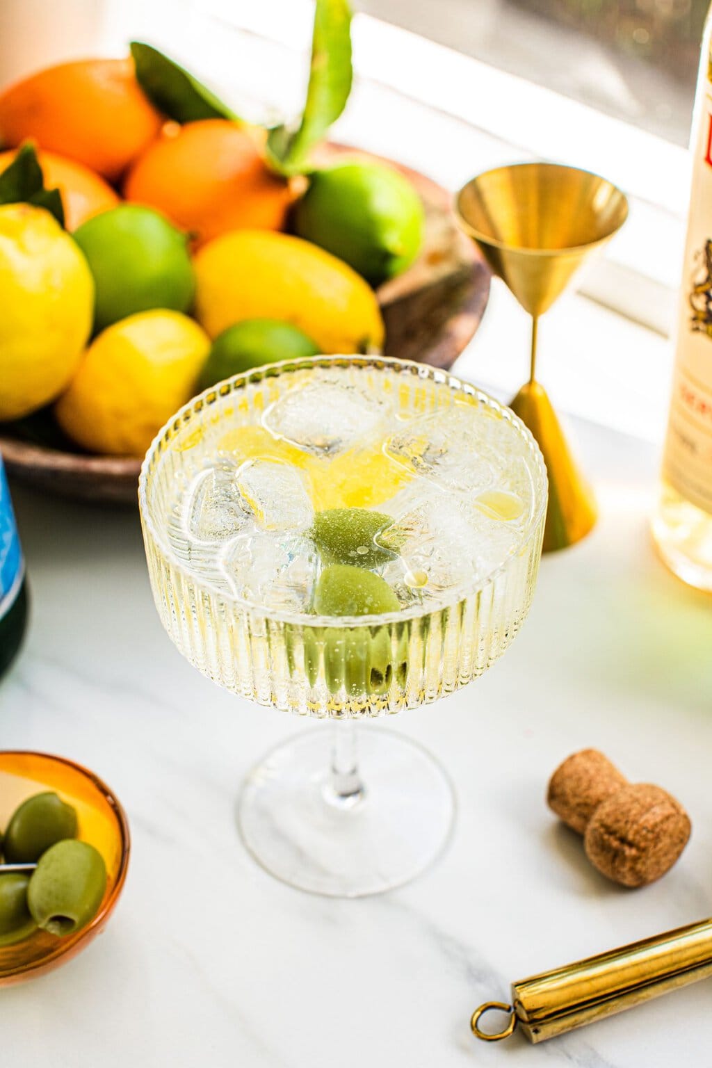 a spritz with olives and lemon inside it and a bowl of oranges, limes, and lemon in the background