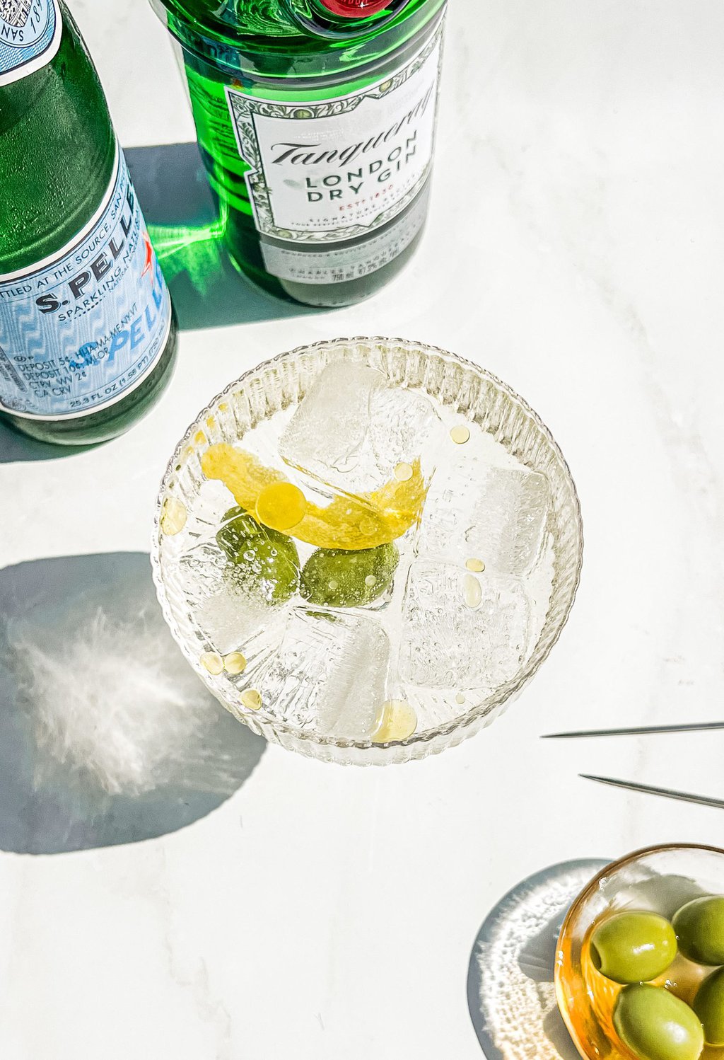 an overhead photo of a martini spritz with lemon rind and olives inside and gin and pellegrino to the side 